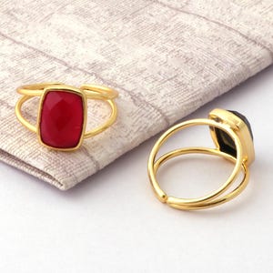 Può includere: Due anelli dorati con pietre rettangolari. Uno presenta una pietra rosso vivo, l'altro una verde scuro. Gli anelli sono appoggiati su un tessuto chiaro e strutturato.