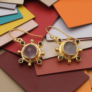 Puede incluir: Un par de pendientes de oro con una piedra preciosa rosa y detalles negros. Los pendientes cuelgan de un gancho y tienen un diseño circular.