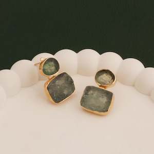 May include: A pair of gold-toned earrings with a two-stone design. Each earring features a small, round, green stone above a larger, rectangular, gray-green stone. The earrings are displayed on a white, scalloped surface against a dark green background.