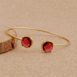 Può includere: Un bracciale aperto color oro con due pietre rotonde sfaccettate di colore rosso intenso, fissate con griffe a ciascuna estremità del sottile cinturino in metallo lucido. Il design è semplice e simmetrico, mettendo in risalto il contrasto.