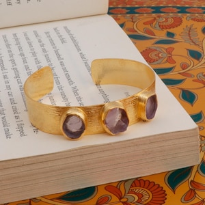 Pode incluir: Um bracelete de ouro com três pedras preciosas roxas engastadas no metal. O bracelete está sobre um livro com um padrão floral.
