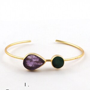 May include: Gold bangle bracelet featuring a raw amethyst and emerald gemstone. The amethyst is teardrop shaped and the emerald is round.