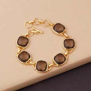 May include: A gold-toned chain bracelet featuring five square-cut, faceted smoky brown gemstones. The bracelet is constructed with circular metal links connecting each stone and includes a T-bar toggle clasp mechanism for secure fastening.