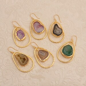 May include: Six pairs of gold-coloured, teardrop-shaped earrings. Each earring features a natural stone in various colours, including purple, pink, black, brown, and green. The stones are set within a double-loop gold frame, creating a unique jewellery design.