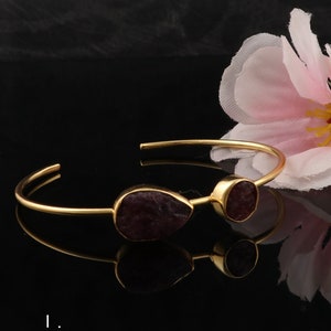 May include: Gold bangle bracelet with two raw ruby stones. The stones are a deep red color and have a natural, unpolished look. The bracelet is simple and elegant.