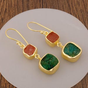 Könnte beinhalten: Ein Paar goldfarbene Ohrhänger mit jeweils zwei quadratischen Steinen. Der obere Stein ist orange und strukturiert, der untere Stein ist grün und strukturiert. Die Ohrringe hängen an goldfarbenen Haken mit einer polierten Metalloberfläche.