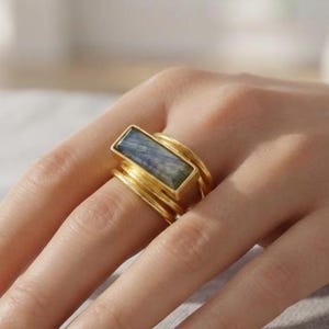 Puede incluir: Un anillo de tono dorado con una piedra de labradorita rectangular e iridiscente montada horizontalmente. La banda está formada por varios hilos finos de color dorado superpuestos que se envuelven alrededor del dedo.