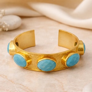 May include: A gold-coloured cuff bracelet featuring five oval, light blue gemstones. The bracelet has a textured surface and an open design. The gemstones are evenly spaced along the front.