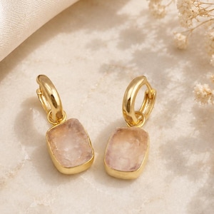 Può includere: Un paio di orecchini a cerchio dorati con pietre preziose rettangolari rosa chiaro. Gli orecchini sono esposti su una superficie di marmo, con un tessuto color crema e fiori secchi sullo sfondo. Gioielli.