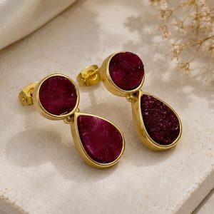 May include: A pair of gold-toned stud earrings featuring two deep magenta, textured druzy stones each. The design consists of a circular stone positioned above a larger teardrop-shaped stone, both encased in smooth, polished gold-colored metal settings.