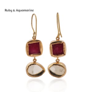 May include: A pair of gold-colored drop earrings. Each earring features a square, ruby-colored gemstone at the top and a clear, aquamarine-colored gemstone at the bottom. The text "Ruby & Aquamarine" is in the upper left corner.