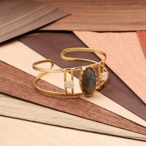 Può includere: Un braccialetto rigido in oro con una grande pietra ovale di labradorite e due pietre bianche più piccole. Il braccialetto è su uno sfondo di legno.