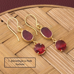 Pode incluir: Brincos dourados com design de duas pedras. Cada brinco apresenta uma pedra fúcsia rosa ovalada em bruto sobre uma pedra vermelha redonda e facetada. Os brincos são exibidos em uma superfície tecida marrom claro. Texto: 1. ASmethyst & Pink Fuchsia.