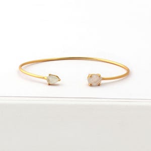 Könnte beinhalten: Ein goldenes Armreif-Armband mit zwei weißen Edelstein-Akzenten. Das Armband ist offen und hat ein schlichtes Design.