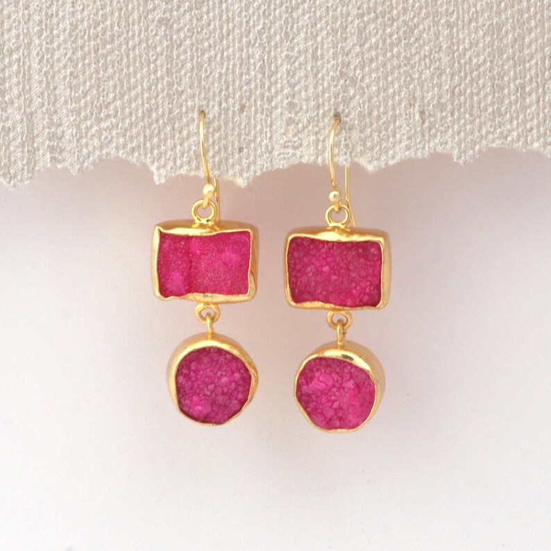 May include: A pair of gold-coloured earrings. Each earring has a rectangular top and a round bottom, both with a vibrant pink, textured stone set in a gold frame. The earrings hang against a white background.