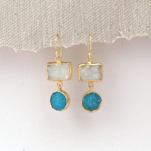 May include: A pair of gold-coloured earrings. Each earring has a rectangular, white stone above a round, turquoise stone. The stones are set in gold-coloured metal. The earrings hang from a simple hook.