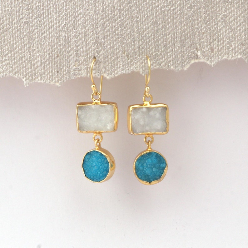 May include: A pair of gold-coloured earrings. Each earring has a rectangular, white stone above a round, turquoise stone. The stones are set in gold-coloured metal. The earrings hang from a simple hook.