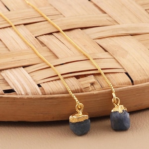 Può includere: Un paio di orecchini a catena dorati con un ciondolo di pietra preziosa blu scuro. Gli orecchini sono appesi a una superficie intrecciata.