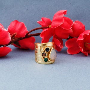 Puede incluir: Un anillo de oro con una banda tejida y una piedra preciosa de ópalo en forma de lágrima. El anillo está adornado con piedras preciosas más pequeñas en tonos de verde, azul y marrón.