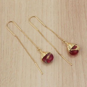 Puede incluir: Un par de pendientes colgantes dorados. Cada pendiente presenta una pequeña piedra roja y dorada envuelta en alambre dorado. Los pendientes se muestran sobre una superficie de madera marrón claro.