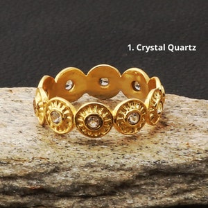 Puede incluir: Un anillo dorado con un patrón repetitivo de diseños circulares, cada uno con un cristal transparente central. El anillo está etiquetado como "1. Crystal Quartz" y descansa sobre una superficie de piedra gris texturizada.