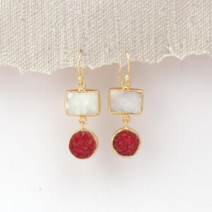 May include: A pair of gold-coloured earrings. Each earring has a rectangular white stone above a round, red stone. The earrings hang from gold-coloured hooks. The background is a neutral, off-white colour.