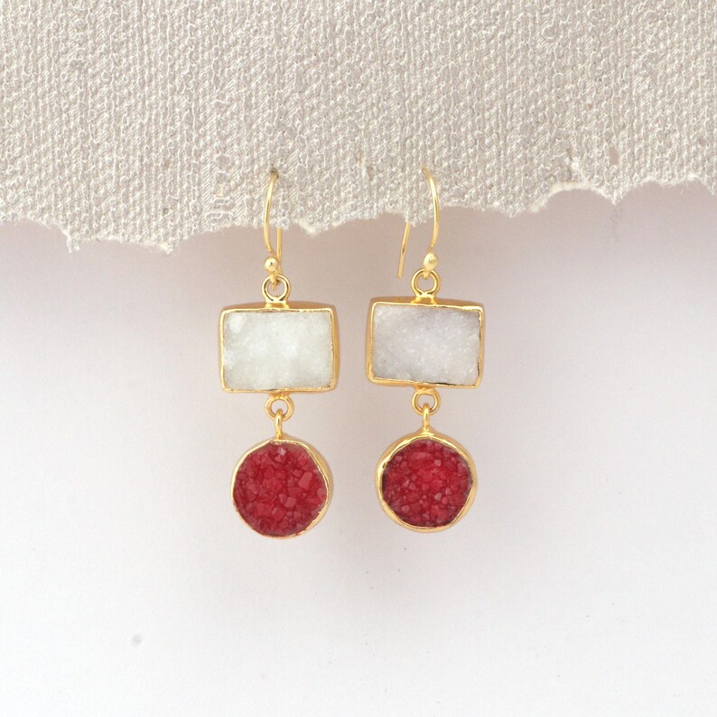 May include: A pair of gold-coloured earrings. Each earring has a rectangular white stone above a round, red stone. The earrings hang from gold-coloured hooks. The background is a neutral, off-white colour.