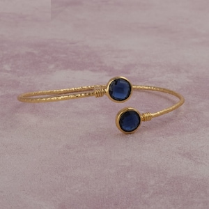 May include: Gold-toned cuff bracelet with a hammered texture. The open design features two round, bezel-set, deep blue gemstones. The bracelet is displayed on a soft, pinkish-purple surface.