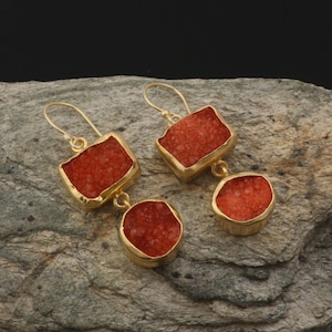 May include: Gold earrings with two red, square-shaped and round-shaped stones. The stones have a textured, sparkly surface.
