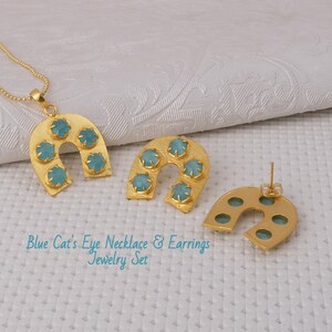 May include: A gold-toned jewelry set featuring a necklace and earrings. The necklace has a pendant with a horseshoe shape, adorned with five light blue gemstones. The matching earrings also have the same design. The set is displayed on a white surface.