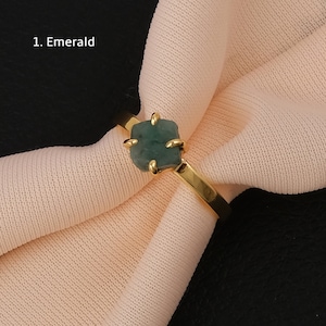 May include: A gold ring with a green emerald gemstone. The ring is on a light pink fabric background. 1. Emerald