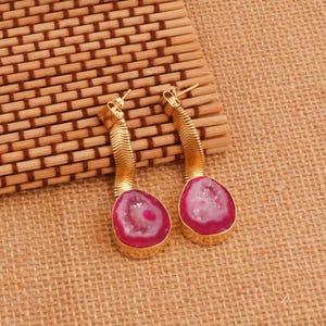 May include: Gold-coloured earrings with a textured, snake-like design. Each earring has a vibrant pink and white stone in a teardrop shape, set in a gold bezel. The earrings are displayed on a woven bamboo mat.