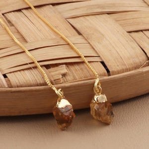 Op de afbeelding: Een paar gouden kettingoorbellen met ruwe, bruine, kristalvormige bedels.