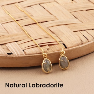 Op de afbeelding: Een paar goudkleurige oorhangers met druppelvormige labradorietstenen. De oorbellen worden gepresenteerd op een geweven, lichtbruin oppervlak. De tekst "Natural Labradorite" staat onderaan de afbeelding.