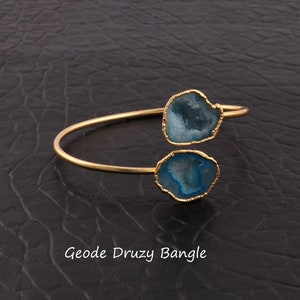 May include: A gold-colored bangle bracelet featuring two blue geode druzy stones with gold-colored edges. The bracelet has a simple, open design. The text "Geode Druzy Bangle" is at the bottom of the image.