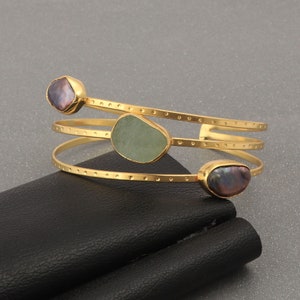 May include: Gold-toned cuff bracelet with three bands. The bracelet features three irregularly shaped stones: two dark purple and one light green. The bracelet is resting on a black surface against a gray background.