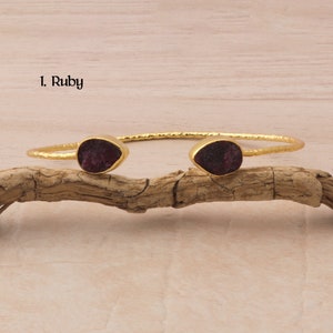 May include: Gold bangle bracelet with two ruby gemstone accents. The bracelet is on a wooden surface. 1. Ruby
