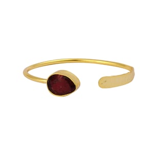 May include: A gold bangle bracelet with a single, dark red gemstone. The bracelet is open and has a hammered texture.