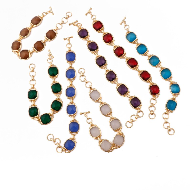 Op de afbeelding: Zeven gouden armbanden met verschillende vierkante edelstenen in verschillende kleuren. De armbanden zijn met elkaar verbonden door een gouden ketting.