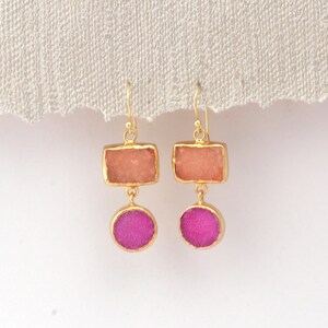 May include: A pair of gold-coloured earrings. Each earring has a rectangular peach-coloured stone above a round, magenta-coloured stone. The stones are set in gold-coloured metal with a simple hook.