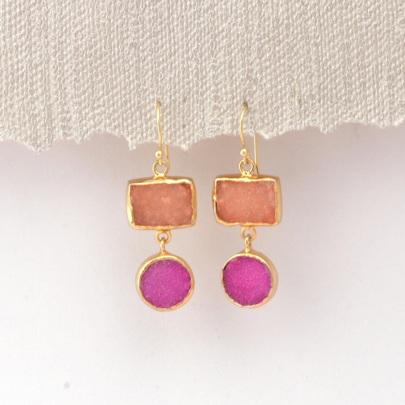 May include: A pair of gold-coloured earrings. Each earring has a rectangular peach-coloured stone above a round, magenta-coloured stone. The stones are set in gold-coloured metal with a simple hook.