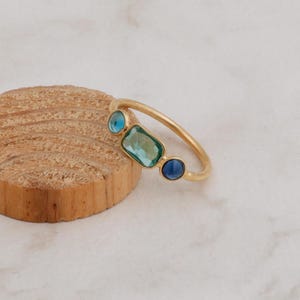 Puede incluir: Un anillo de oro con tres piedras preciosas azules y verdes. El anillo está sobre una superficie de madera.