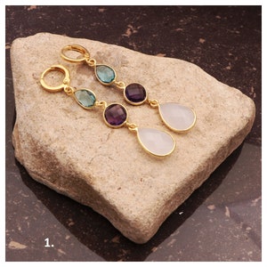 May include: Gold dangle earrings with three teardrop-shaped gemstones. The top gemstone is a light blue colour, the middle gemstone is a dark purple colour, and the bottom gemstone is a white colour.