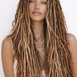 May include: A woman with long, layered dreadlocks in shades of brown and blonde. The hair frames the face, with a fringe across the forehead. She wears a cream-colored top and multiple layered necklaces.