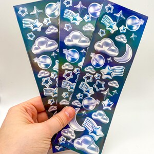 May include: Three sheets of iridescent blue holographic stickers with a space theme. The stickers feature stars, moons, clouds, and planets.