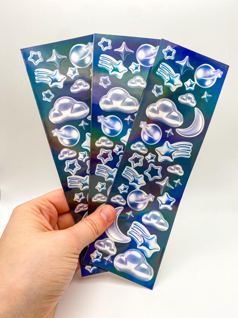 May include: Three sheets of iridescent blue holographic stickers with a space theme. The stickers feature stars, moons, clouds, and planets.
