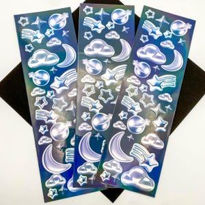 May include: Three sheets of iridescent holographic stickers featuring a variety of space-themed designs, including stars, moons, clouds, and planets. The stickers are on a black background.