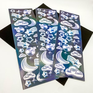 May include: Three sheets of iridescent holographic stickers featuring a variety of celestial designs, including moons, stars, and clouds. The stickers are perfect for adding a touch of whimsy to journals, planners, and other craft projects.
