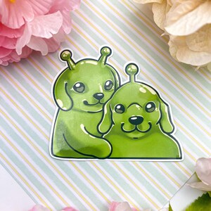 May include: A sticker featuring two green alien dogs with big eyes and cute expressions. The dogs are hugging each other and have a glossy finish.
