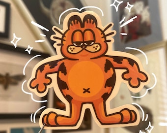 May include: A die-cut sticker featuring Garfield, the orange cartoon cat, with a grumpy expression. The sticker has black spots and a white outline with motion lines and starbursts. A pair of tweezers is holding the sticker.